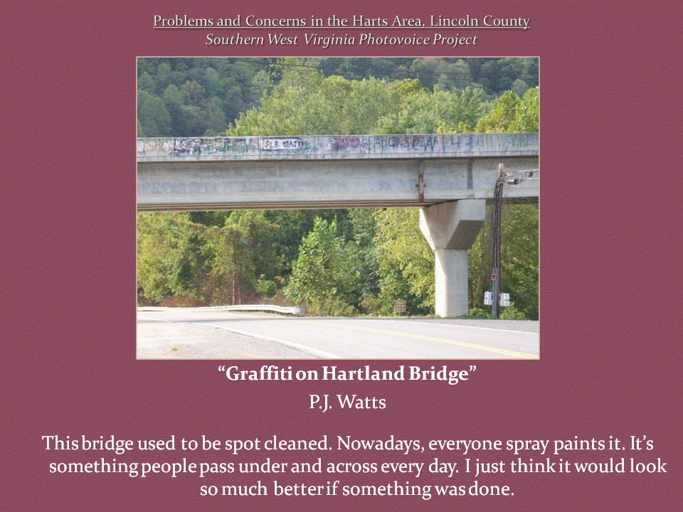 Graffiti covers the side of a concrete highway overpass supported by a large pillar above a road surrounded by wooded hills.