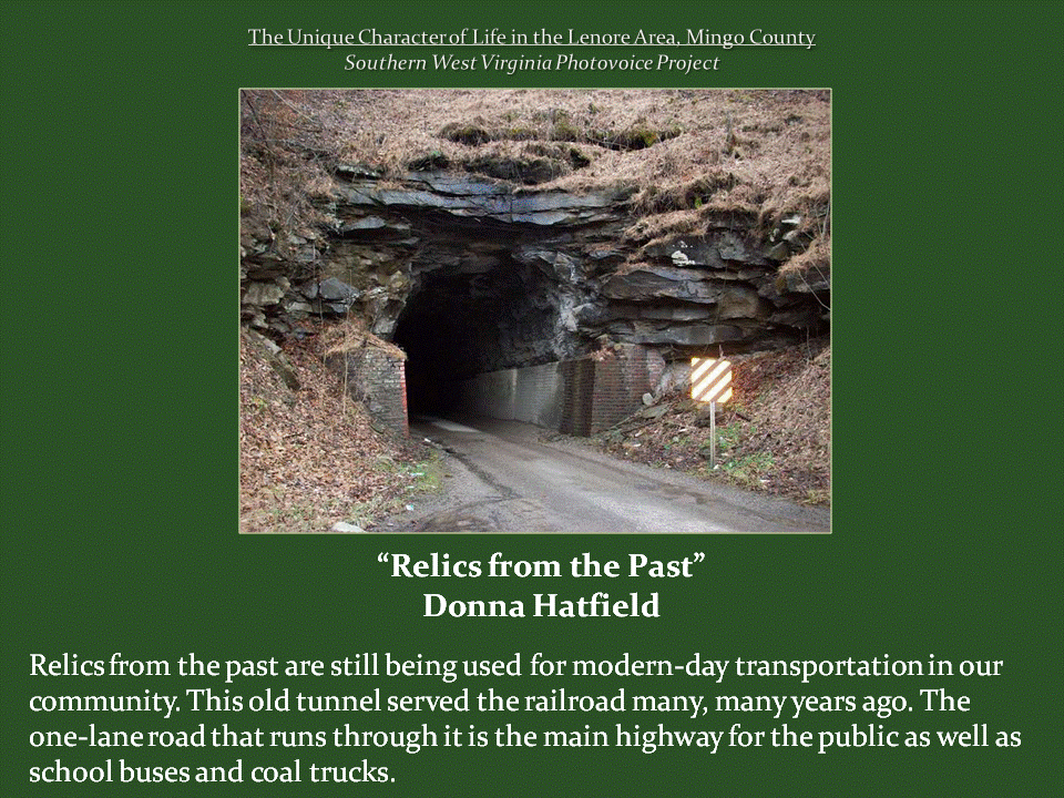 A narrow paved road enters an old railroad tunnel carved through a rocky hillside, with brick supports framing the entrance and a striped roadside warning sign. The historic tunnel now serves as a one-lane highway used by local traffic, school buses, and coal trucks.