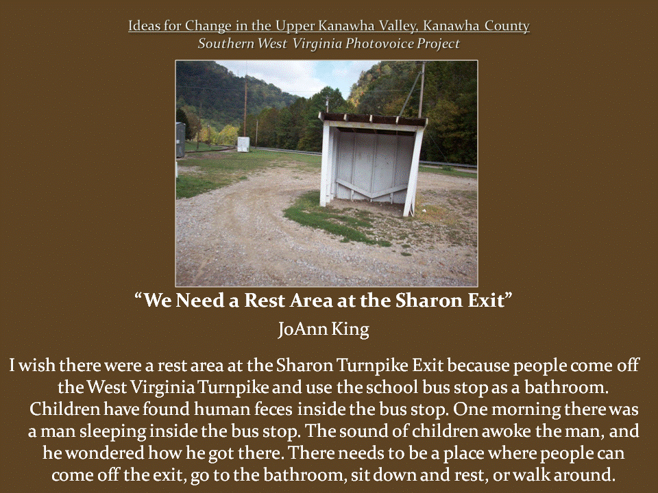 Small, three-sided school bus shelter with a slanted roof stands beside a gravel turnaround near a wooded mountain road.