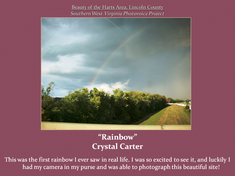 A faint rainbow arcs across a cloudy sky above green trees and a grassy hillside.