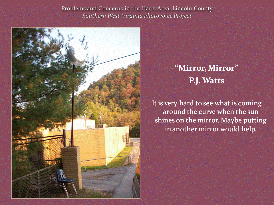 A convex traffic mirror mounted on a pole beside a building, with trees and a wooded hillside in the background.