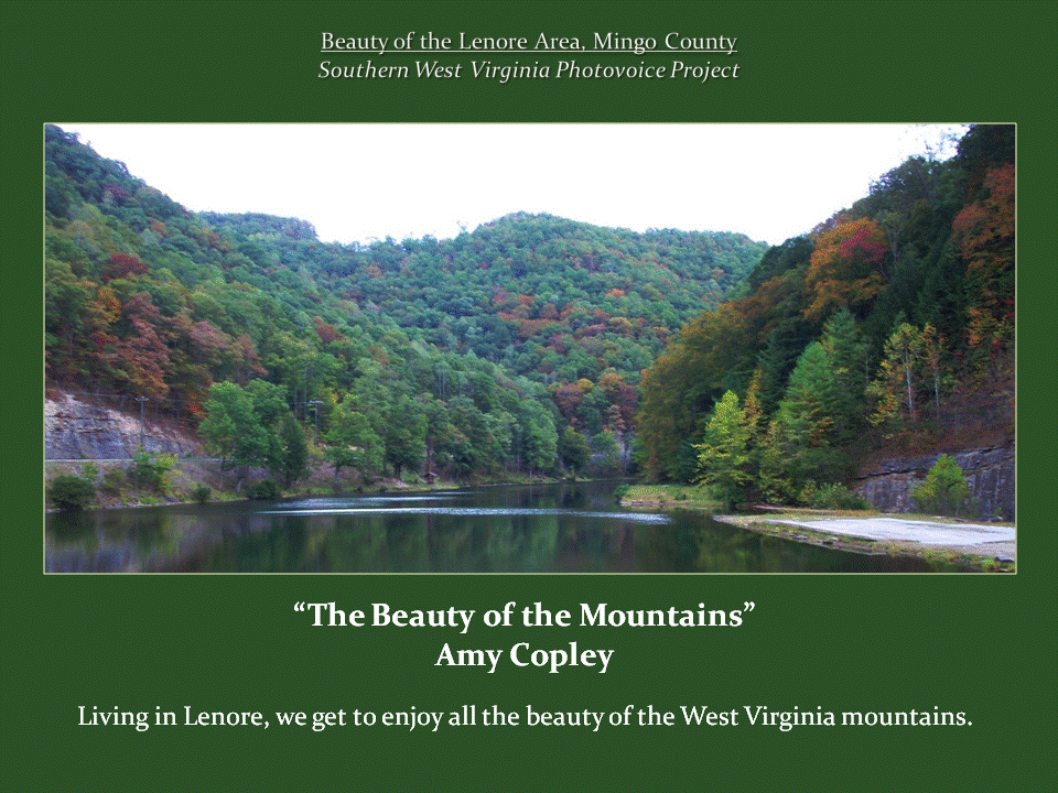 A peaceful lake surrounded by densely forested mountains displaying green, yellow, and red autumn foliage, with a narrow road following the shoreline.