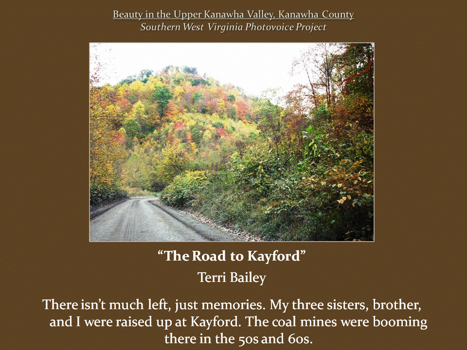 Narrow rural road curves through dense vegetation toward a mountainside covered with trees displaying vibrant autumn foliage.