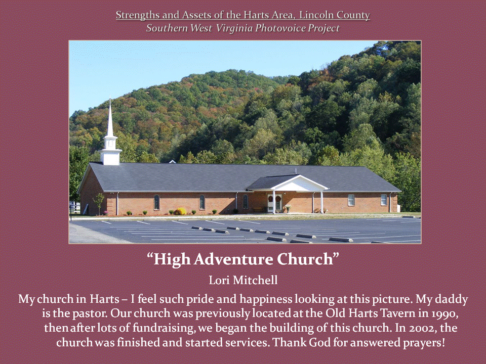 A long brick church with a white steeple stands before a wooded hillside, with an empty parking lot in front.