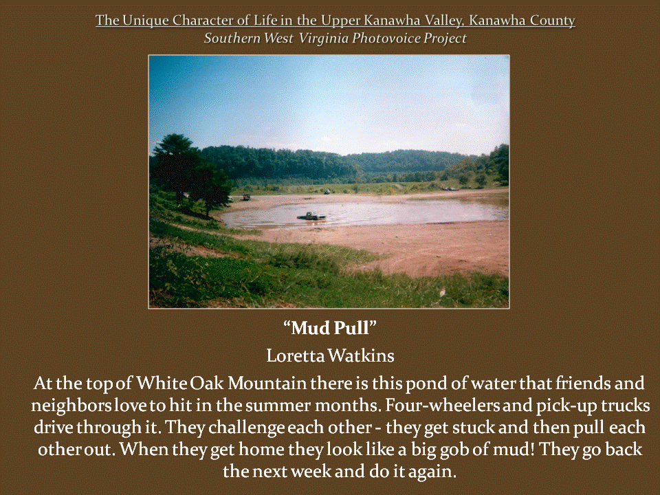 Wide, muddy pond in a mountain clearing, with vehicles crossing shallow water and wooded hills surrounding the open area.