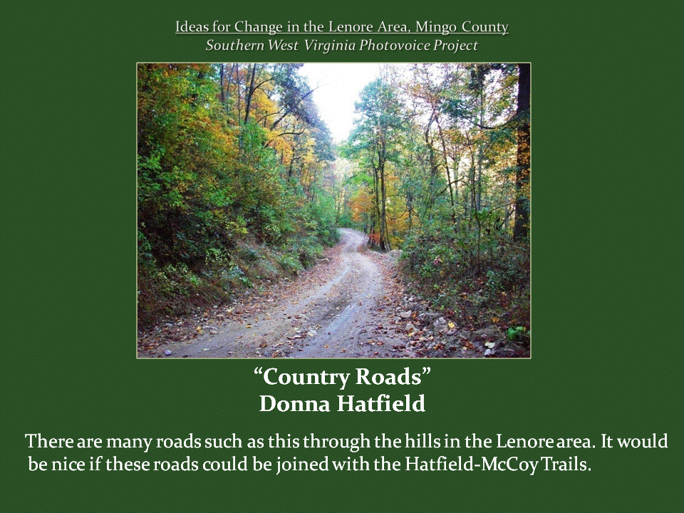 A narrow, unpaved country road winds through dense woodland with green and yellow autumn foliage.