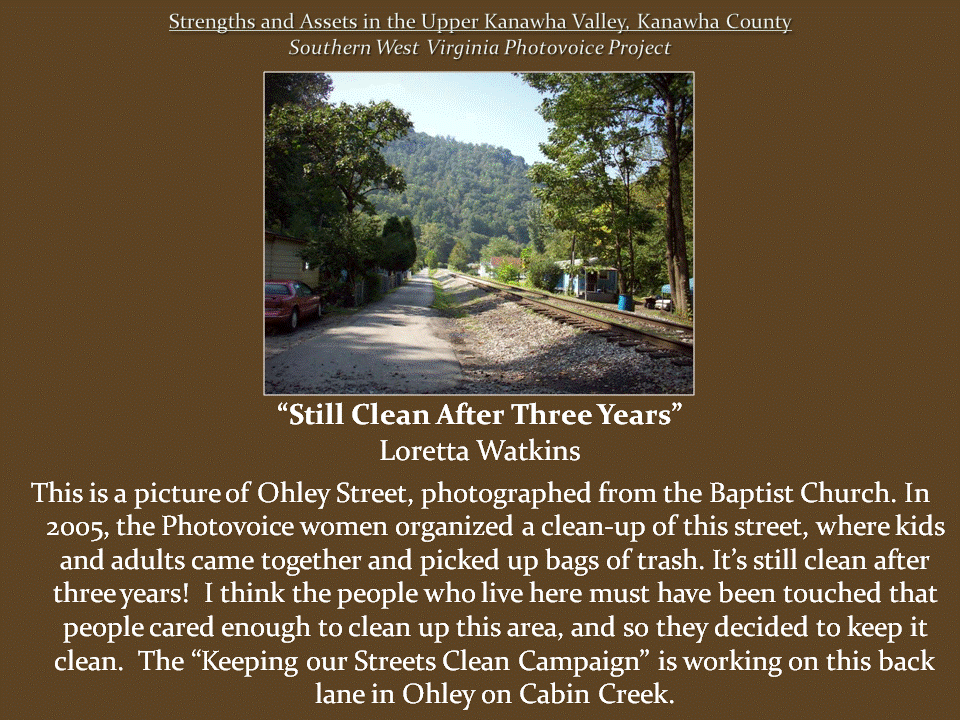 Clean residential lane runs alongside railroad tracks, with houses, trees, and a forested mountain in the background.